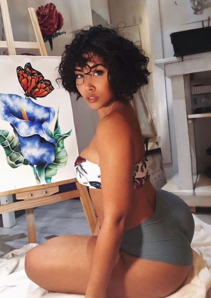 Artistic FemDom companion with short curly hairstyle Confident woman posing beside floral artwork Premium fetish phone chat profile image Elegant dominant female companion portrait Creative fantasy chat companion profile Stylish FemDom profile photograph Premium adult chatline companion image Curvy artistic companion portrait Modern confident woman with natural curls