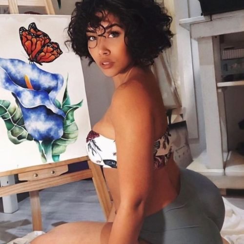 Artistic FemDom companion with short curly hairstyle Confident woman posing beside floral artwork Premium fetish phone chat profile image Elegant dominant female companion portrait Creative fantasy chat companion profile Stylish FemDom profile photograph Premium adult chatline companion image Curvy artistic companion portrait Modern confident woman with natural curls