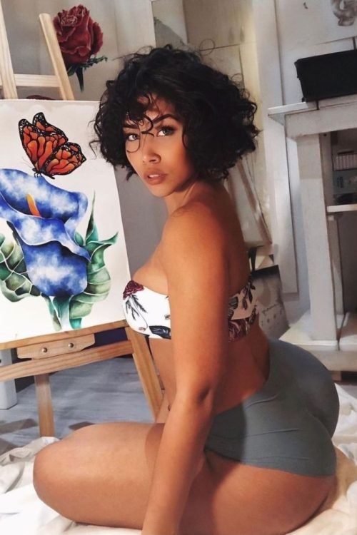 Artistic FemDom companion with short curly hairstyle Confident woman posing beside floral artwork Premium fetish phone chat profile image Elegant dominant female companion portrait Creative fantasy chat companion profile Stylish FemDom profile photograph Premium adult chatline companion image Curvy artistic companion portrait Modern confident woman with natural curls